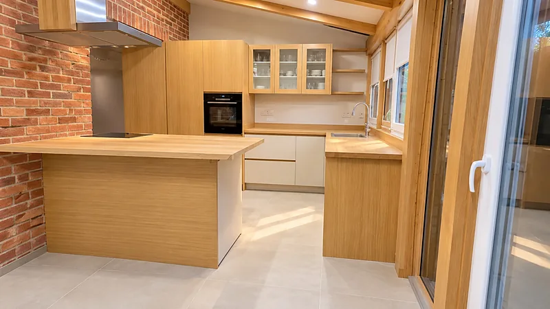 Cuisine en bois clair avec îlot central et mur de briques, intégrée dans une véranda