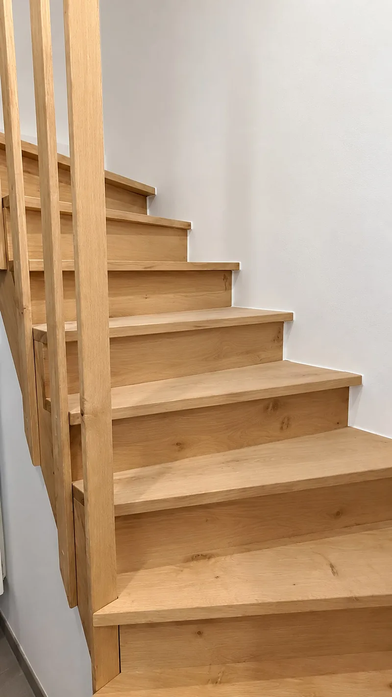 Volée d'escalier en bois clair avec garde-corps à barreaux, le long d'un mur blanc