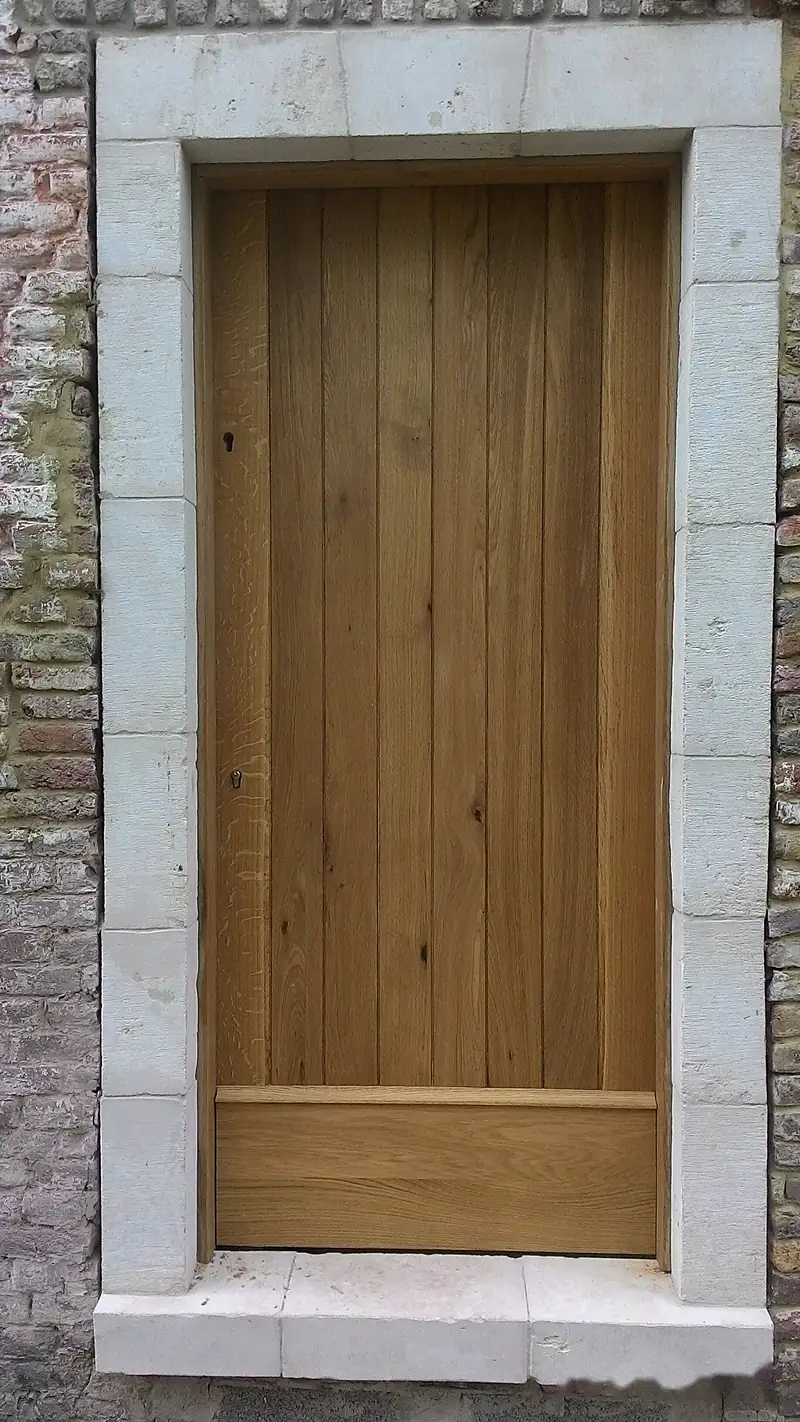 Porte extérieure en bois clair à planches verticales, dans un encadrement en pierre